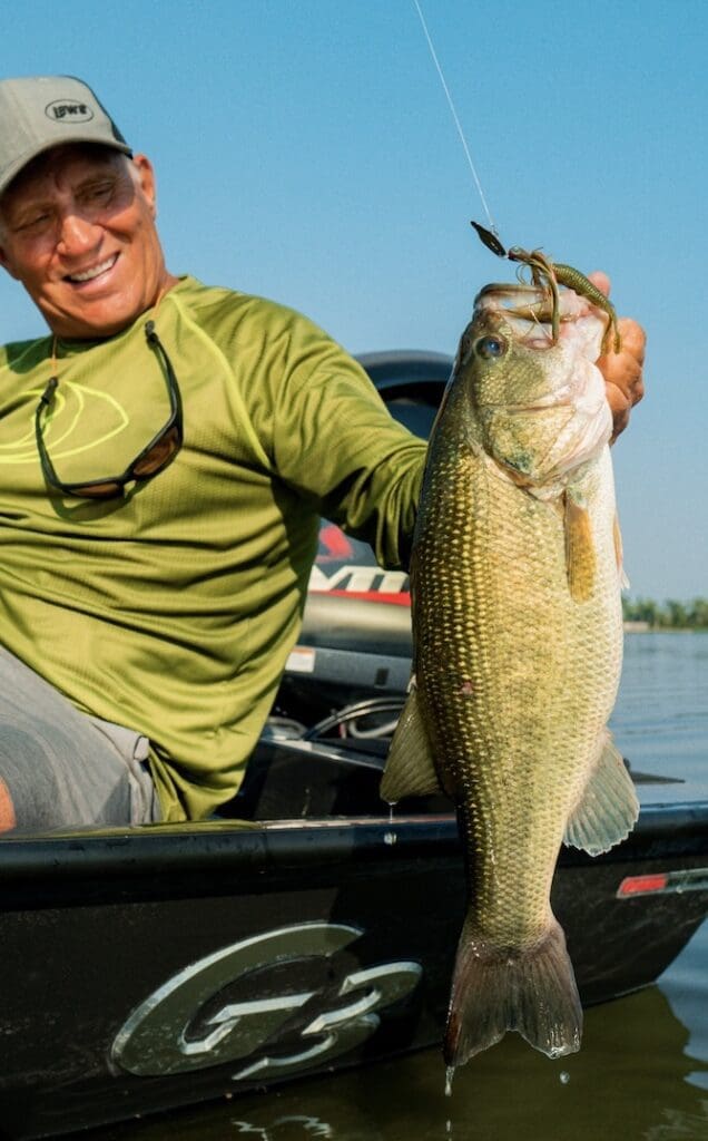 A smiling man in a green shirt and cap sits on a boat, holding up a large bass fish he caught while fishing the Midwest, with blue sky and water in the background.