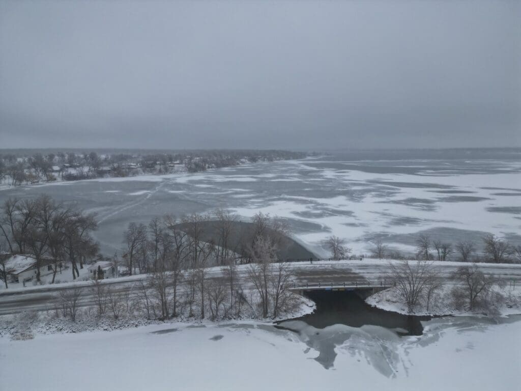 A wintry landscape featuring a partially frozen lake with shimmering ice, snow-covered ground, bare trees, and a small bridge over a creek leading into the lake beneath a gray, cloudy sky.