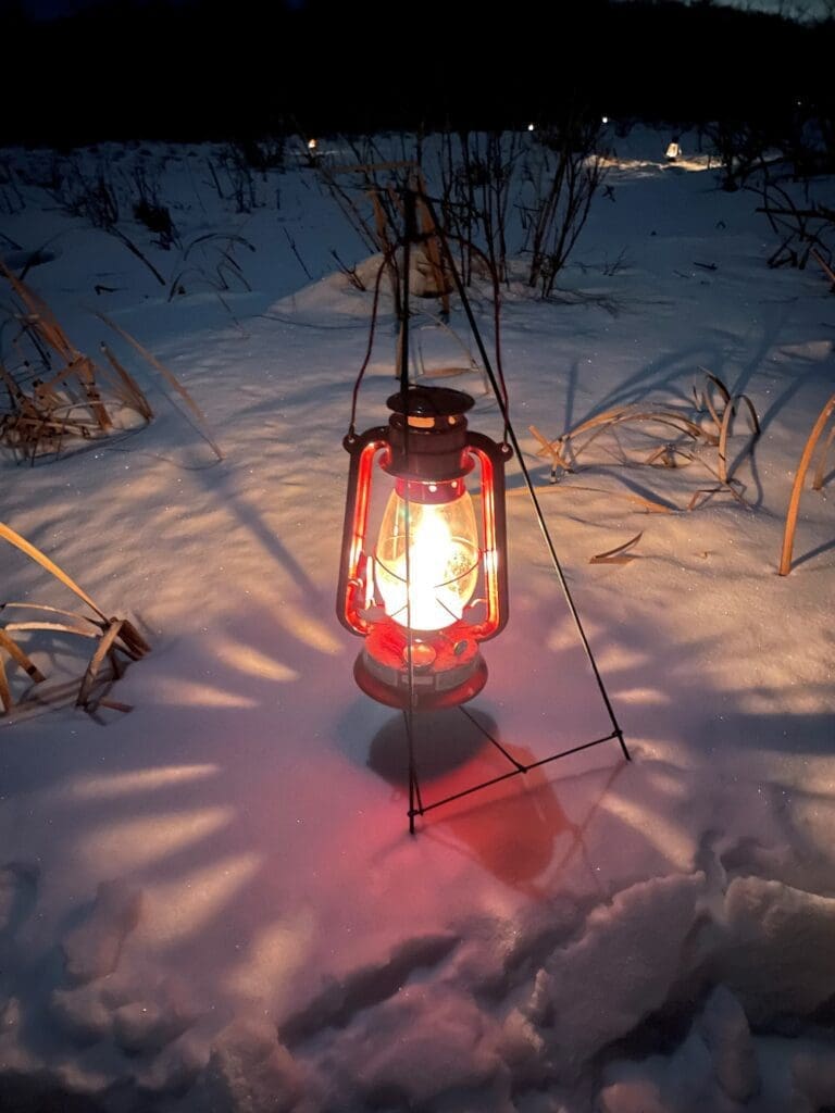 A red lantern glows warmly on snow-covered ground at dusk during a 2022 Candlelit Walk in the park, surrounded by dry grass and a dark outdoor landscape.