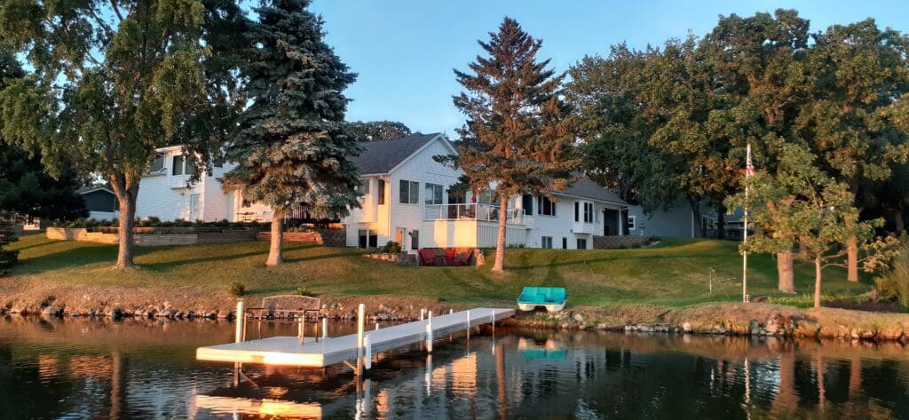 The Cottage on Lake Henry features a white house with large windows on a grassy hill by the lake, surrounded by trees. A small dock extends over the water as sunset light glows on the house, lawn, and docked rowboat.