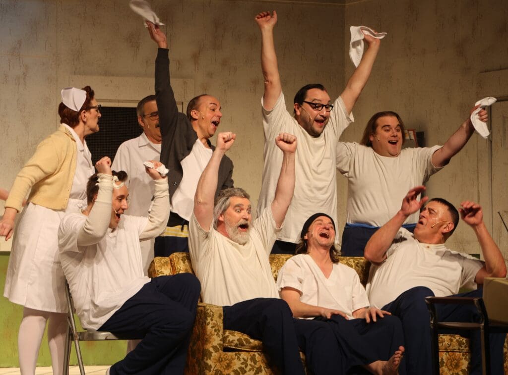 A group of people dressed as nurses and patients cheer enthusiastically, raising their arms and waving handkerchiefs, while sitting and standing around a couch in a lively, joyful scene at the Andria Theatre.