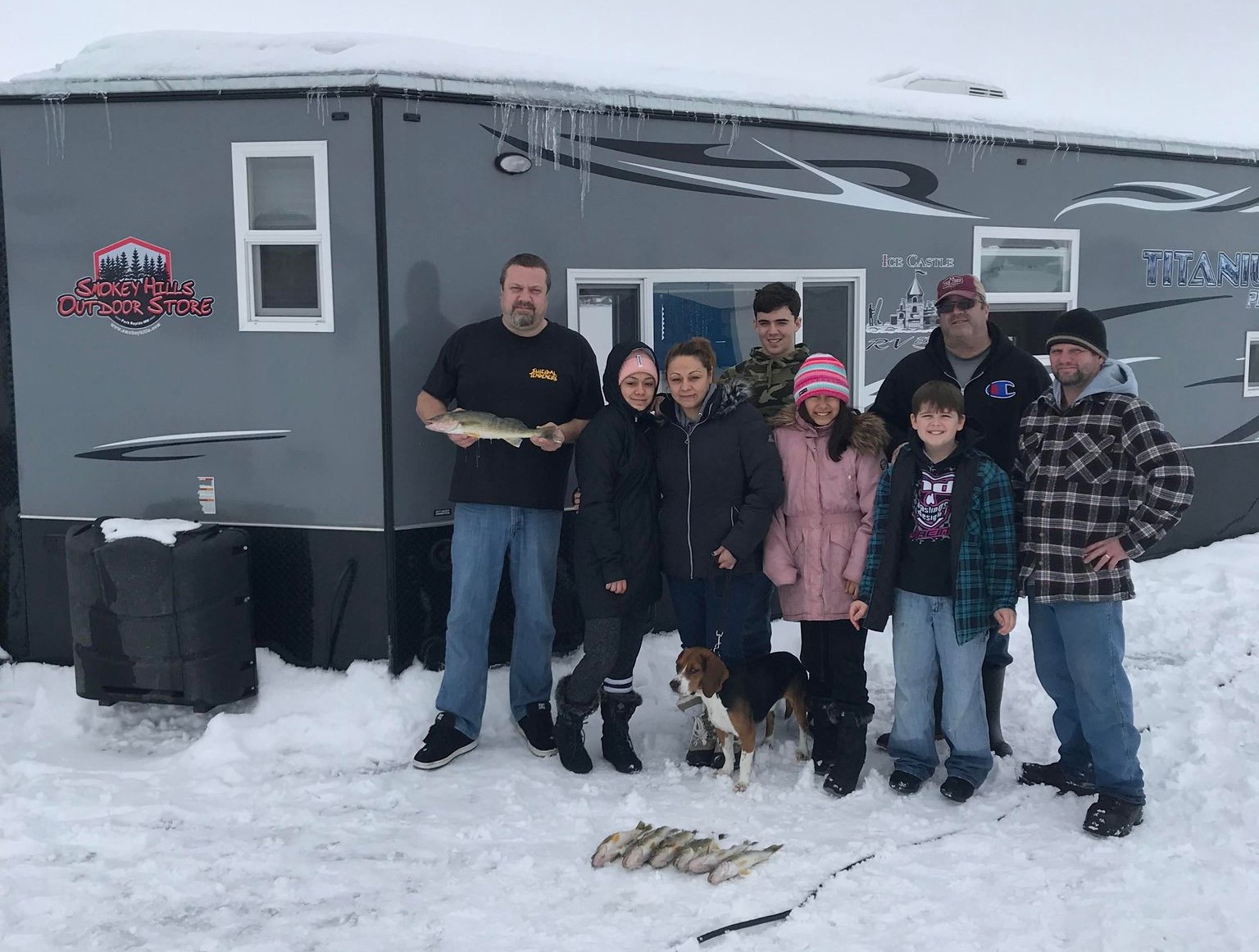 Eight people and a dog pose in front of a dark gray Tyler Wolden Fishing ice fishing trailer on snow, with several fish laid out in front. The group is warmly dressed for winter.