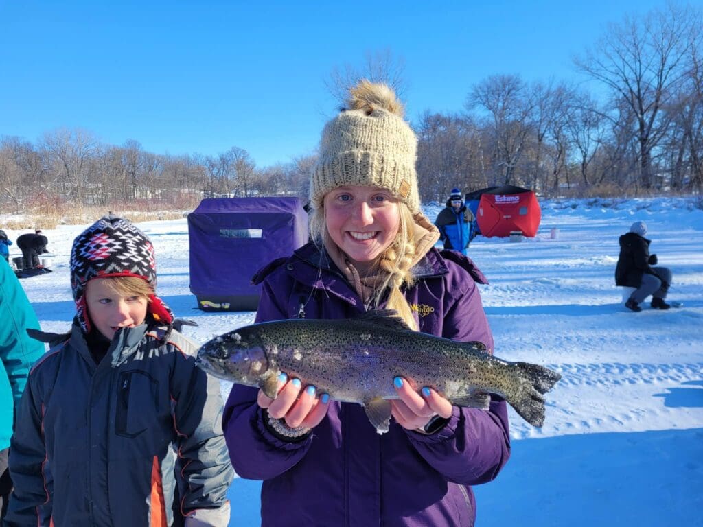 A smiling woman in a knit hat and purple coat holds a large fish on a snowy, frozen lake during an ice fishing trip. A boy stands beside her, with people ice fishing in shelters visible in the background under a clear blue sky.