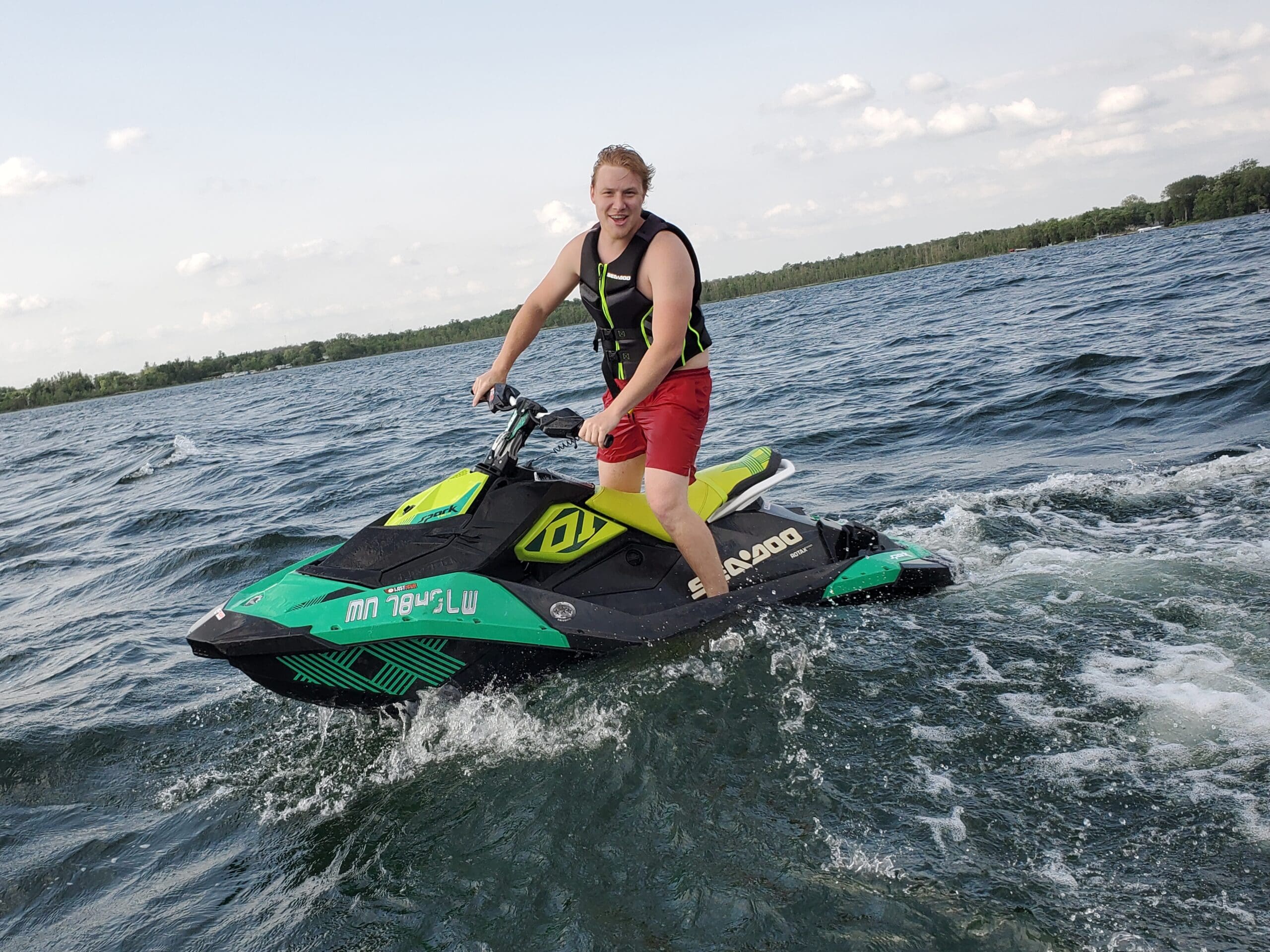 A person wearing a life jacket and red shorts is riding a green and black jet ski from Alexandria Jet Ski Rental on a lake, smiling at the camera with trees and a cloudy sky in the background.