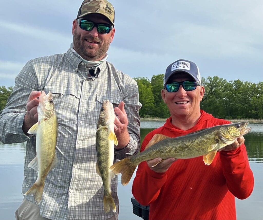Two men in sunglasses and hats stand by a lake, smiling and holding up three fish they caught while fishing the Midwest. Trees and water are visible in the background under a cloudy sky.