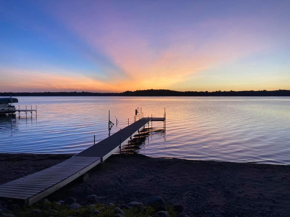 A wooden dock extends over calm lake water at sunset in Sandy Beach Haven, with vibrant orange, yellow, and purple hues lighting up the sky. Trees form a dark silhouette along the distant shoreline.