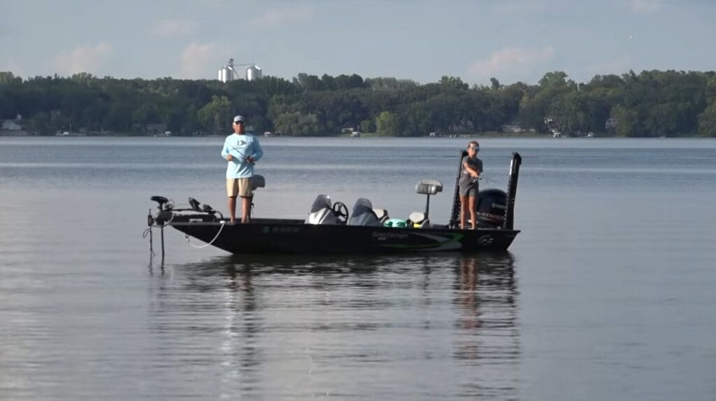 Two people are fishing on a black boat in a calm lake, surrounded by trees and distant buildings under a clear sky—an idyllic scene of fishing the Midwest.