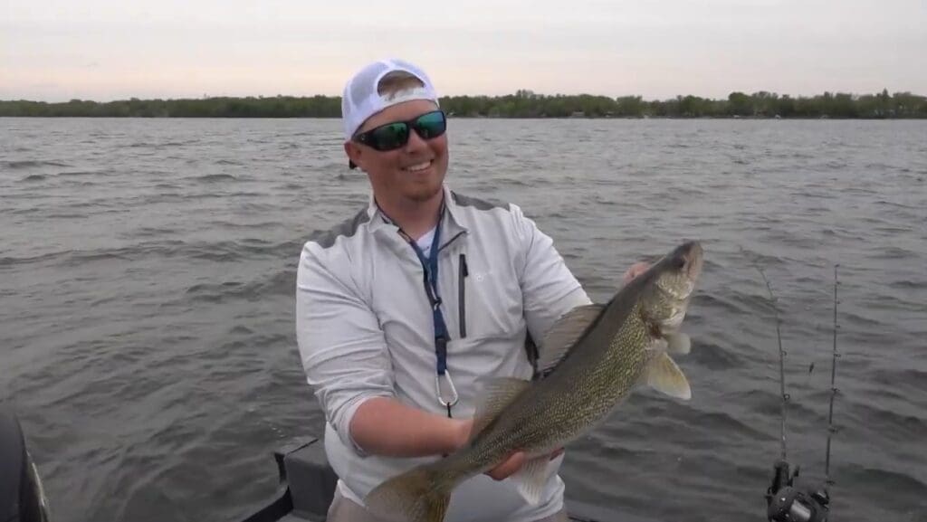 A smiling man wearing sunglasses and a white cap holds a large walleye on a boat in a lake, with shimmering water and distant trees in the background.