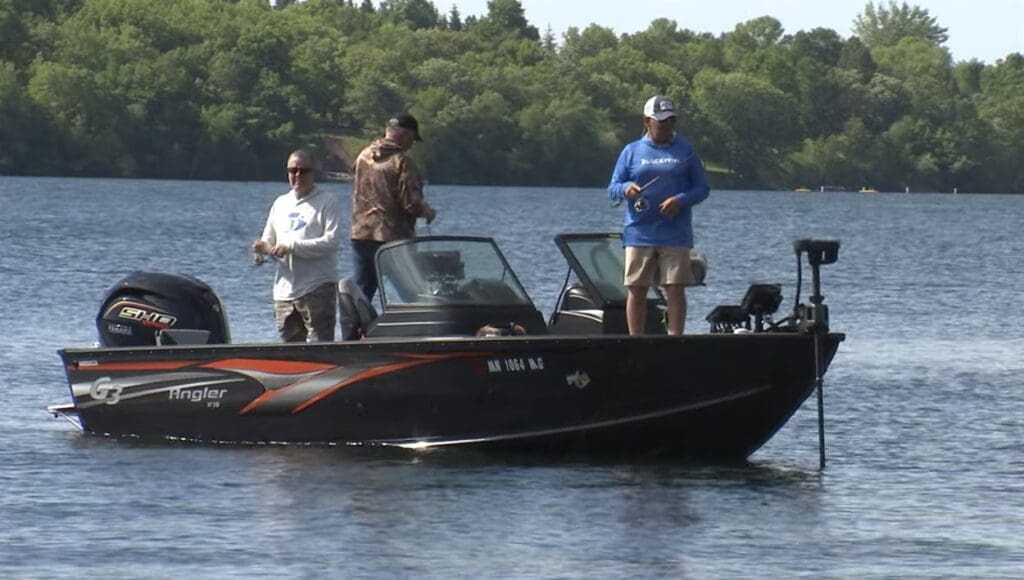 Three people are fishing on a black motorboat with orange stripes on a calm lake, surrounded by lush trees and greenery along the shore.