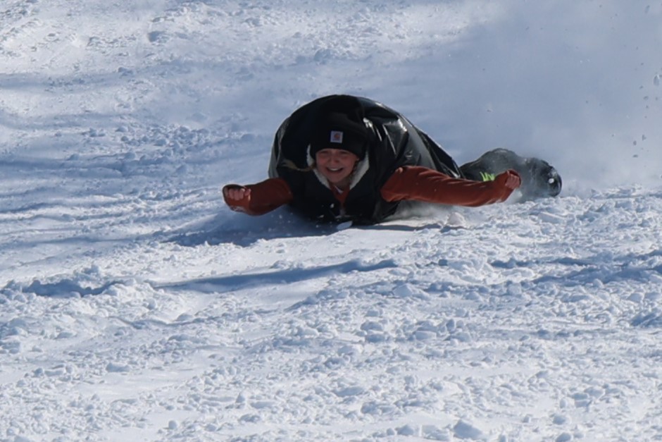 A person wearing a winter hat and gloves slides down a snowy hill face-first on a black sled, smiling as snow kicks up behind them. Bright sunlight highlights the wintry scene—perfect for skiing in Alexandria MN.