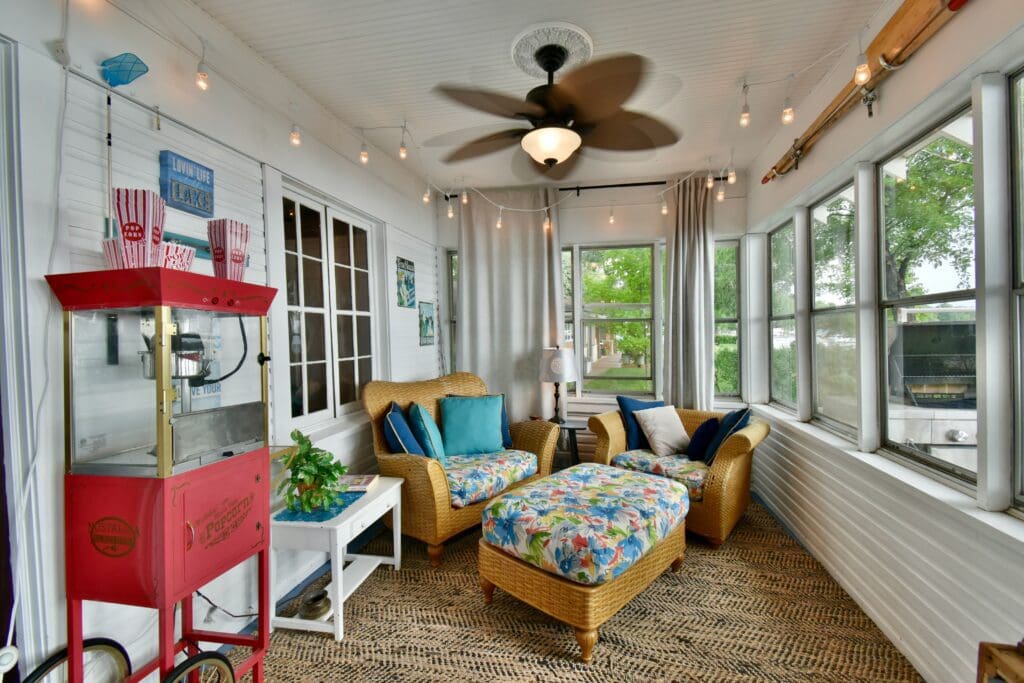 A cozy sunroom at Lake Le Homme Dieu Cottage- Mary's Place features wicker chairs and ottoman with blue cushions, a ceiling fan, string lights, popcorn machine, side table, and large windows with natural light and views—perfect for your Alexandria VRBO stay.