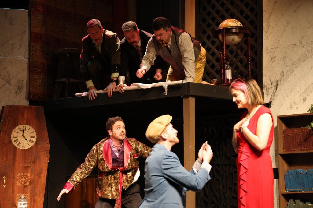 Five men in vintage costumes act dramatically on a two-level stage, with three leaning over a balcony above. Below, one man kneels, holding a ring to a surprised woman in red—an engaging proposal scene for your weekend theatre plans.