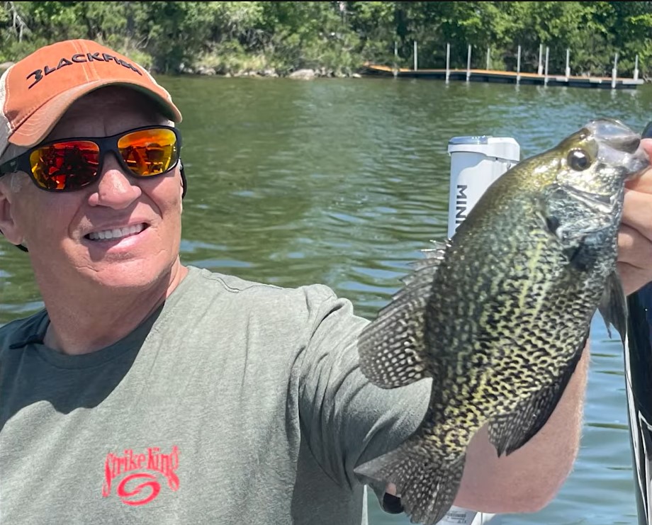 A smiling man in sunglasses and a cap holds up a large crappie fish on a boat, showcasing his success fishing the Midwest, with a lake and trees in the background.