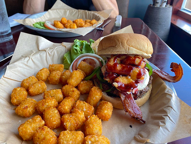 A tray at DJ's Tap House with crispy tater tots, a burger topped with bacon, onion rings, and BBQ sauce, plus lettuce, tomato, and onion on the side. Another similar meal sits in the background on a wooden table.