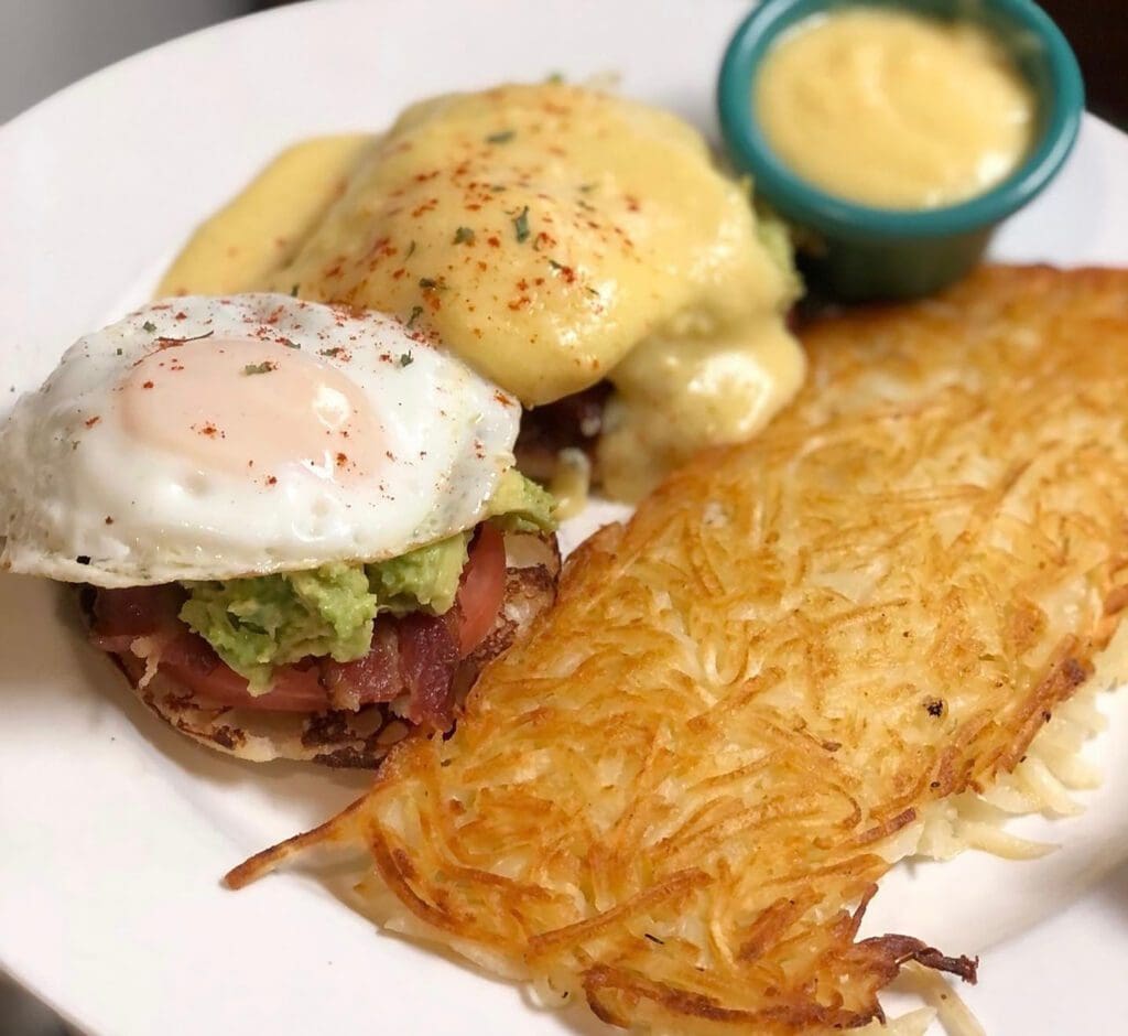 A delicious RESTAURANT WEEK breakfast plate featuring eggs Benedict with hollandaise, a fried egg with avocado and bacon on an English muffin, crispy hash browns, and a small cup of yellow sauce—perfect for breakfasts lovers.