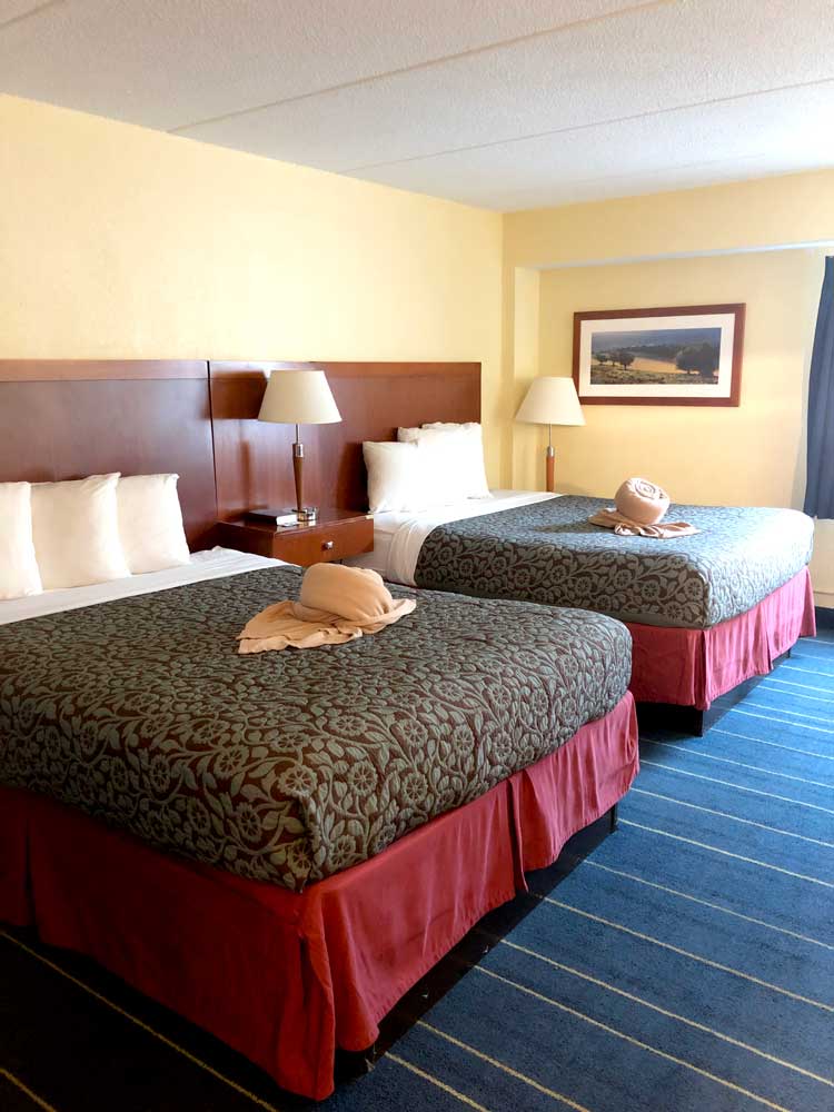 Experience comfort at Relax Inn, one of the top hotels in Alexandria Minnesota. This hotel room features two double beds with patterned bedspreads, towel art, white pillows, bedside tables with lamps, framed artwork, and striped blue carpet.