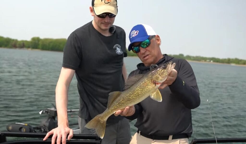 Two men on a boat, one in a black T-shirt and beige cap, the other in sunglasses and a blue cap, proudly hold an Alexandria walleyes catch with greenish-yellow scales over the water. Trees and shoreline can be seen in the background.