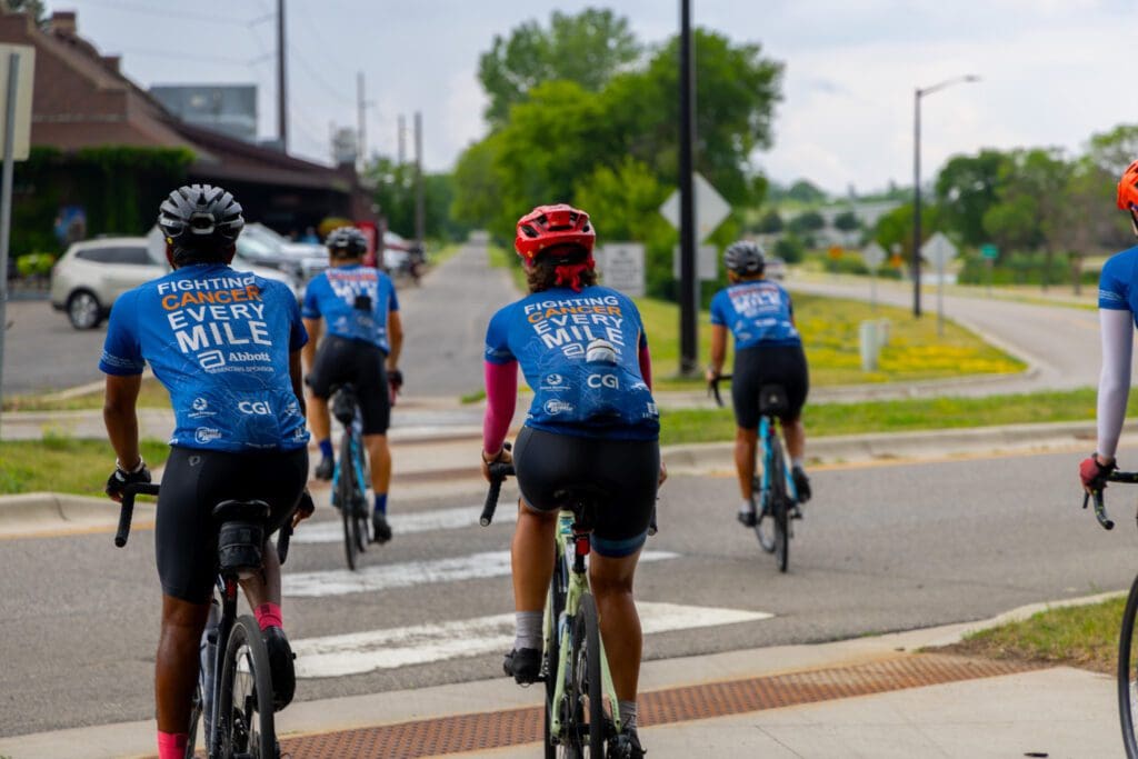 Five cyclists in matching blue jerseys reading "FIGHTING CANCER EVERY MILE" ride together on a cloudy day, heading toward a crosswalk. They're part of the Alexandria to Texas to Alaska bike ride, raising awareness as they journey onward.