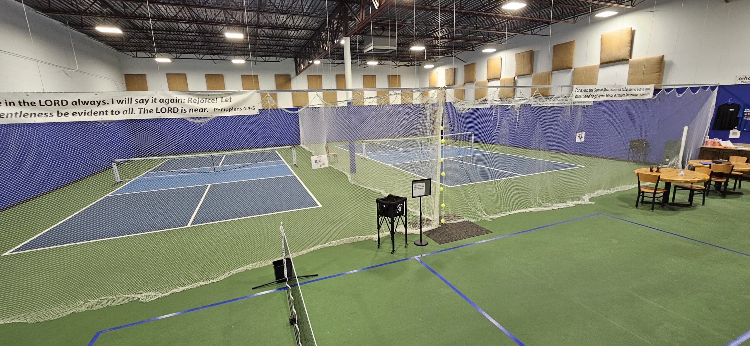 Indoor sports facility featuring multiple pickleball courts at Blind Squirrel Pickleball Club, with green flooring, netting between courts, tables and chairs on the right, and a biblical banner hanging above the courts.