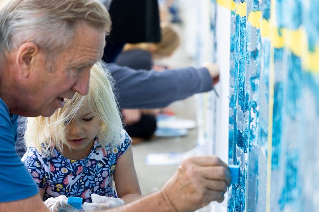 An older man and a young girl with blonde hair paint a mural together outdoors as part of the Alexandria community mural project. Both are focused on applying blue paint to a wall, while others work on the community mural in the background.