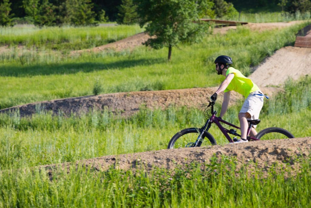 A person wearing a helmet, neon green shirt, and shorts enjoys physical activity as they ride a mountain bike over a dirt trail surrounded by green grass and trees on a sunny day.