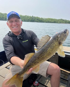 A smiling man sitting in a boat on a lake holds up a large walleye with both hands. Trees and water are visible in the background on a sunny day, capturing the perfect fishing moment in the Alexandria area.