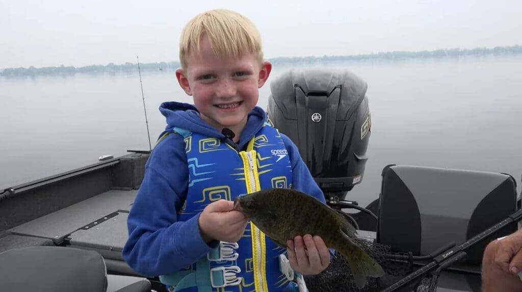 A young boy wearing a blue life jacket smiles while holding a small fish on a boat, enjoying fishing the Midwest with a calm lake and cloudy sky in the background.
