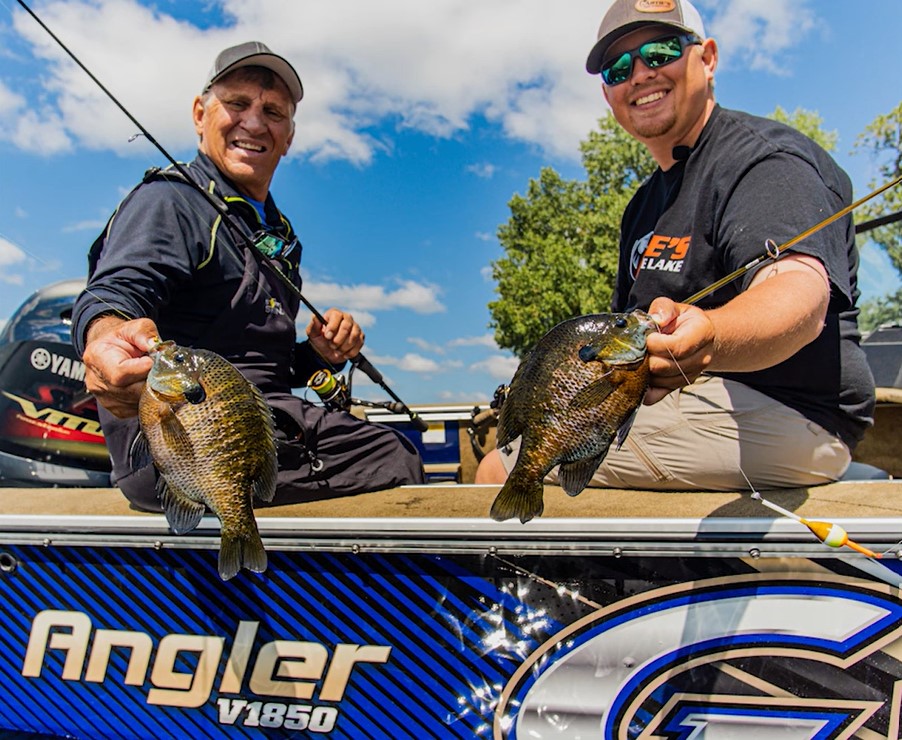 Two smiling men sit in a boat labeled “Angler v1850,” holding up freshly caught fish. Enjoying multi-species fishing in the Alexandria area, they wear sunglasses and caps with blue sky and trees in the background.