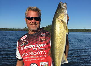 A smiling man wearing sunglasses and a red fishing jersey holds up a large fish in one hand while standing on a boat—showcasing the success of a Minnesota Fishing Guide Service—against a blue lake and tree-lined shore.