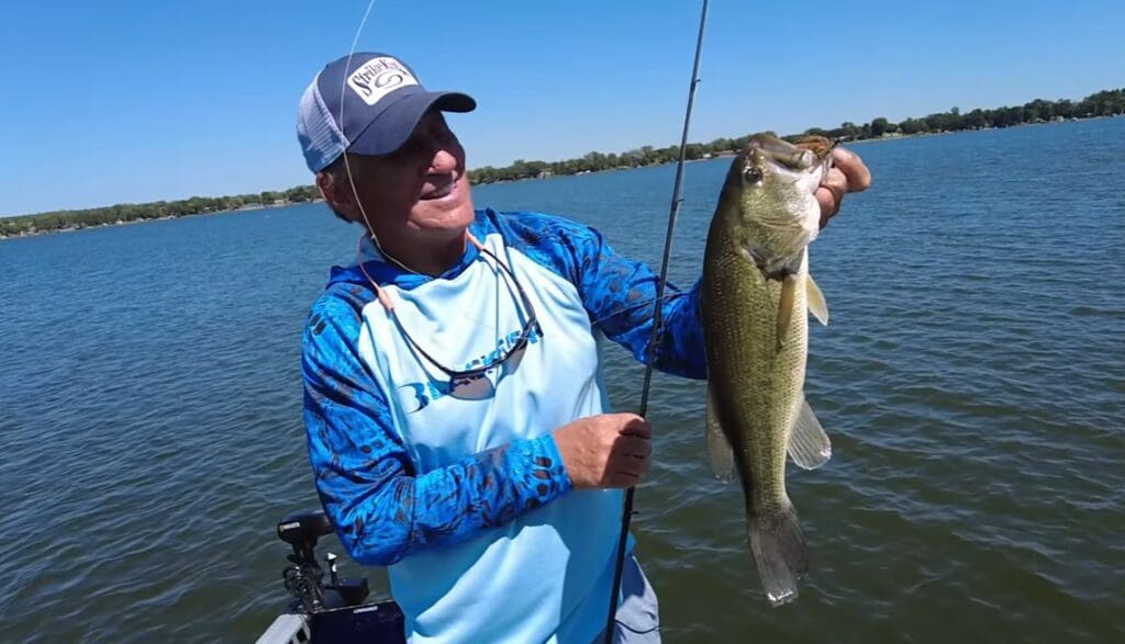 A man in a blue fishing shirt and cap holds up a large fish he caught while fishing the Midwest from his boat on a lake, with clear blue water and a distant tree-lined shore under a sunny sky.