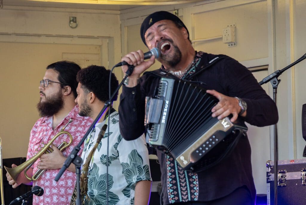 Three musicians perform on stage in Alexandria. One sings passionately into a microphone while playing an accordion, evoking a true Zydeco Experience, while the other two, holding brass instruments, look to the side. All wear patterned shirts.