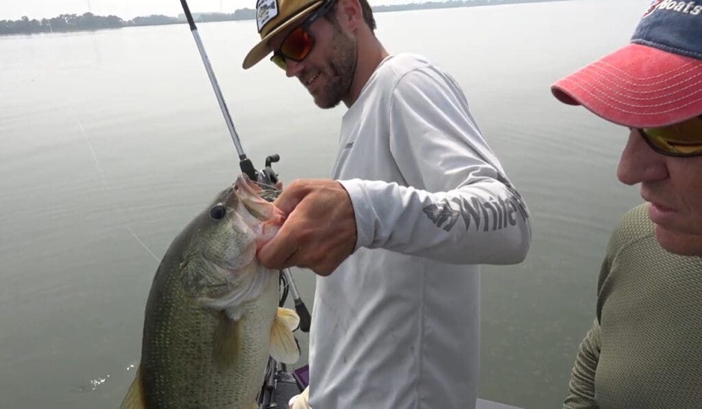 A man in sunglasses and a hat holds a large fish with one hand while standing on a boat, showcasing the thrill of fishing the Midwest. Another person in a red cap stands nearby, with calm water and a hazy shoreline in the background.