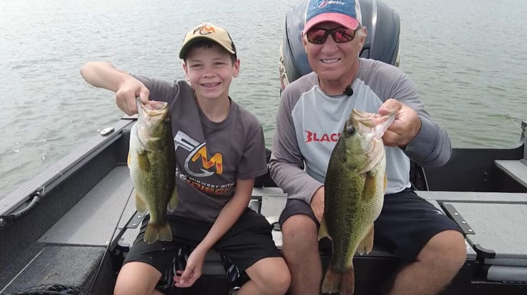 Two people sit in a boat on a lake, smiling and holding up big bass they caught while fishing the Midwest. The younger person wears a cap and T-shirt, while the older one sports sunglasses, a cap, and a long-sleeve shirt.