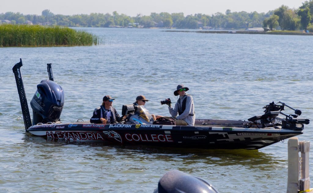 Three fisherman sit in a black Alexandria College fishing boat on a lake, surrounded by water and green shoreline, with fishing equipment visible onboard—ready for their next Champions Tour adventure.