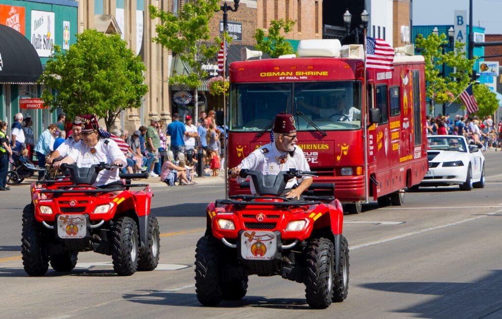 Two Shriners wearing fezzes ride red ATVs in a parade through Alexandria, followed by a red Osman Shrine van. Spectators line the street and American flags are visible in the background, creating a lively Midwest scene.