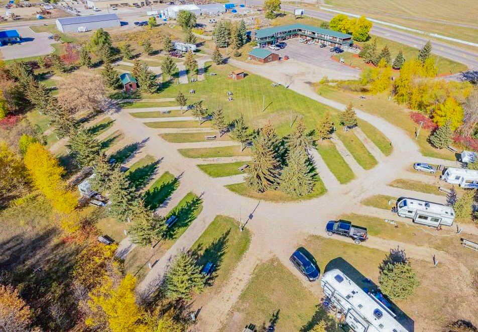 Aerial view of an RV park with several RVs parked on gravel pads among green trees, open grassy areas, and a large building in the background near a road. Some autumn foliage is visible—perfect for your next "RV There Yet" adventure.