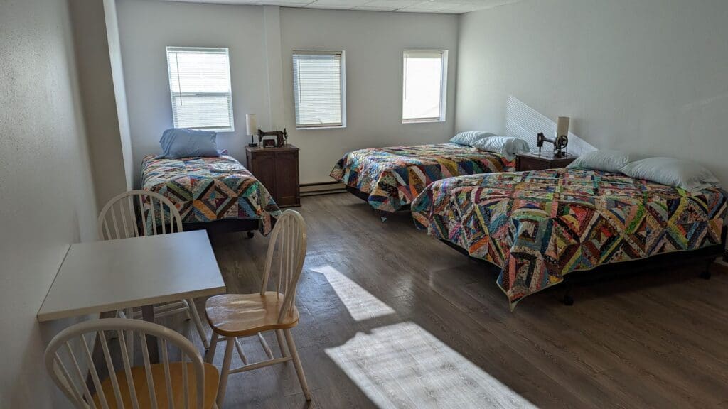 A bright room at Dawn's Quilt Shop & Retreat Center features three beds with colorful patchwork quilts, two nightstands with lamps, a small desk with two chairs, and sunlight streaming through three windows.