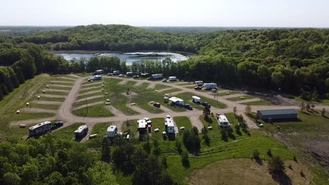 Aerial view of Andes RV Park campground with multiple parked RVs and trailers, winding gravel roads, scattered trees, a building, and a pond surrounded by dense green forest in the background.