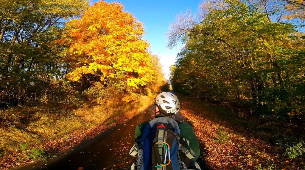 A person biking across Minnesota, wearing a helmet and backpack, cycles along a tree-lined path in autumn, with vibrant yellow and orange foliage under a clear blue sky.