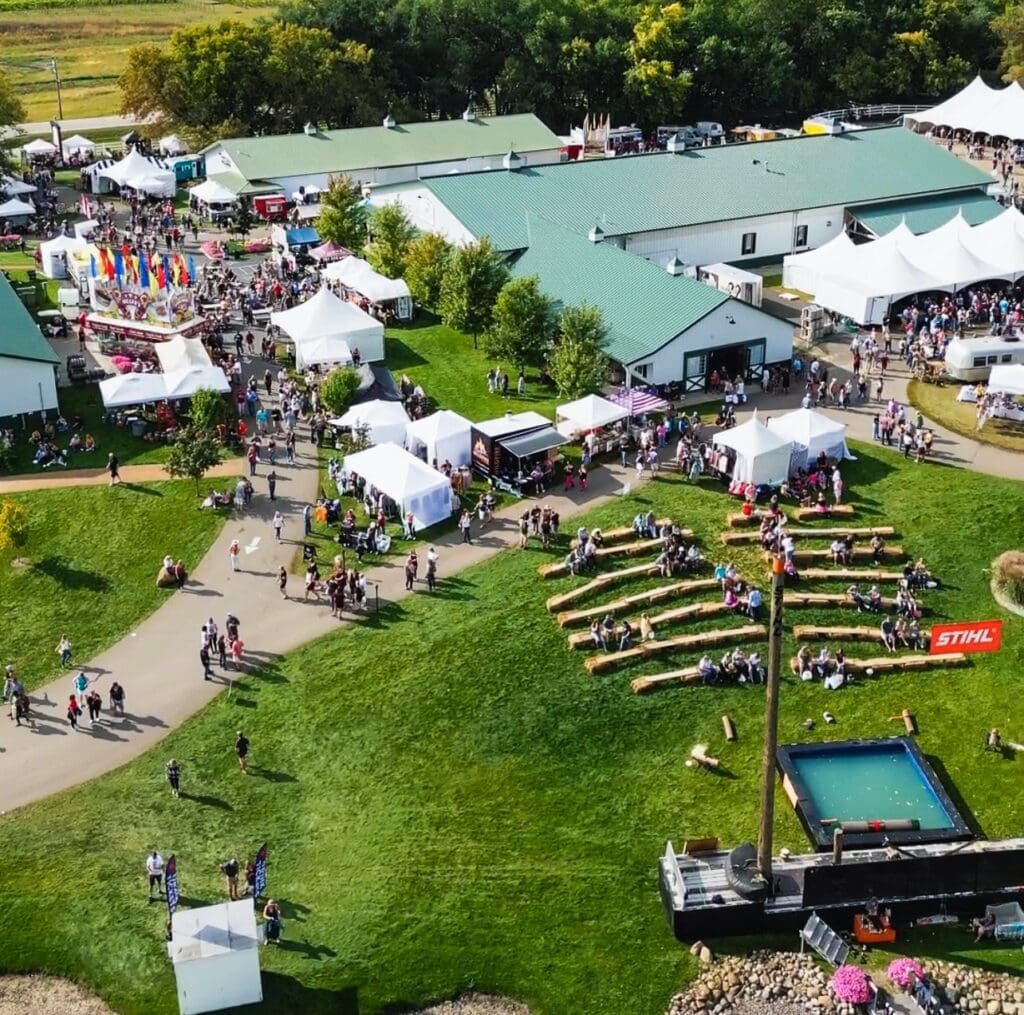 Aerial view of the Grape Stomp Festival at Carlos Creek Winery, featuring tents, vendor booths, walking paths, a green-roofed building, and a STIHL-branded woodcutting demonstration area with logs and a small pool on the grass.