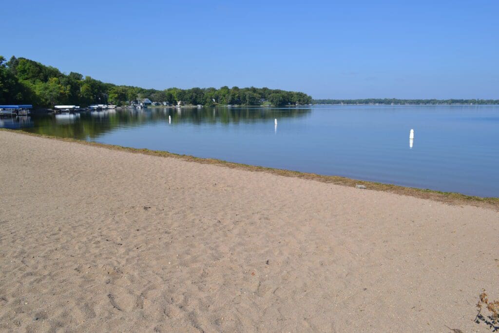 A sandy beach with calm water, a tree-lined shoreline, and a clear blue sky awaits at Lake Le Homme Dieu Beach in Alexandria, MN. White buoys float in the water and a few docks with boats are visible in the distance.