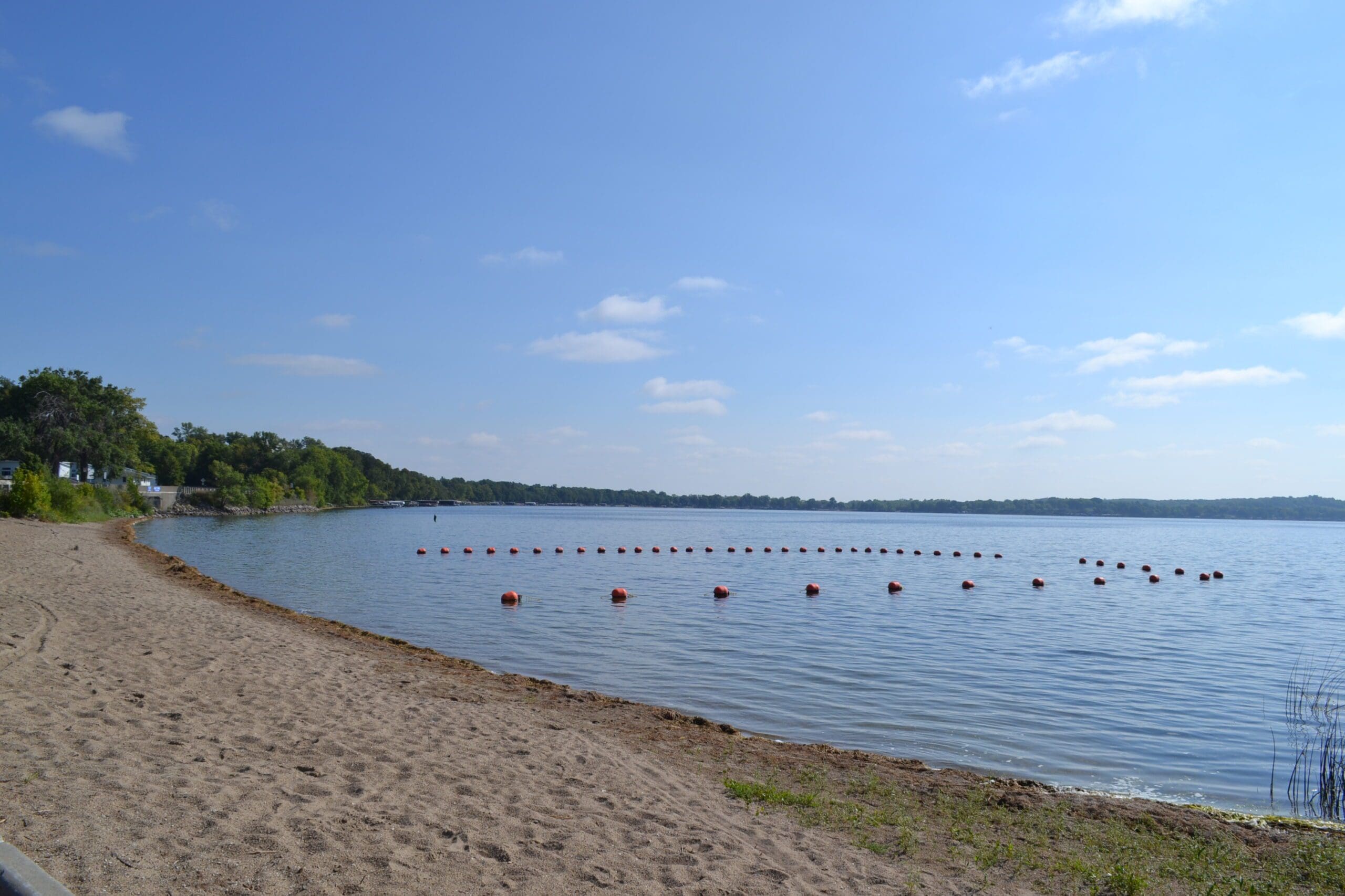 A sandy beach curves along calm lake waters at Rotary Beach Alexandria MN, with a line of red buoys floating under a clear blue sky and green trees surrounding Lake Le Homme Dieu.