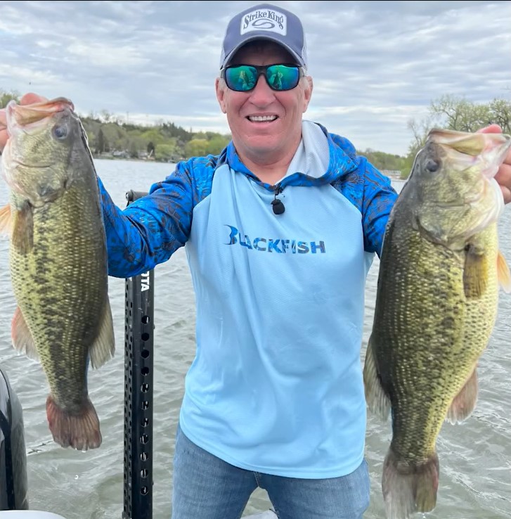 A smiling man in a blue fishing shirt and cap holds up two large bass fish, showcasing his season highlights while standing on a boat in a lake. Trees and water are visible in the background.