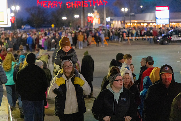A large crowd gathers outdoors at night under a lit "Merry Christmas" sign during one of the lively Christmas events in Alexandria. In the foreground, a woman carries a child on her shoulders while people in winter clothes enjoy the festive, illuminated scene.