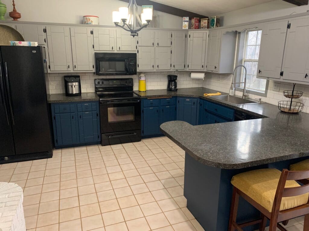 Modern kitchen with blue and gray cabinets, black appliances, a Crafters Edge black countertop, tiled floor, and a breakfast bar with two yellow-cushioned stools. A window above the sink lets in natural light.
