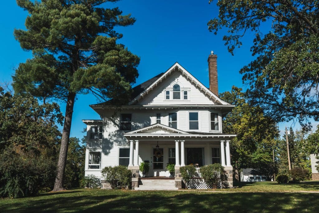 A large, white, two-story house—The Starling House—with a front porch and gabled roof sits on a grassy lawn, surrounded by tall trees under a clear blue sky, offering one of the most charming places to stay in Alexandria MN.