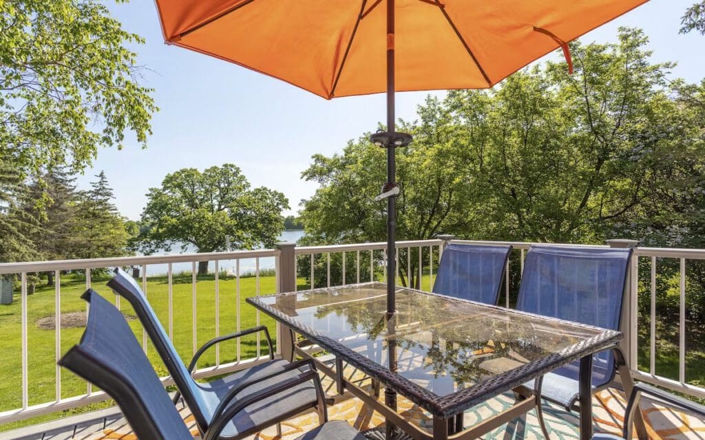 A glass patio table with four blue chairs sits under an orange umbrella on a balcony, perfect for a Taylor-Made Stay, overlooking green trees and a lake in the background on a sunny day.