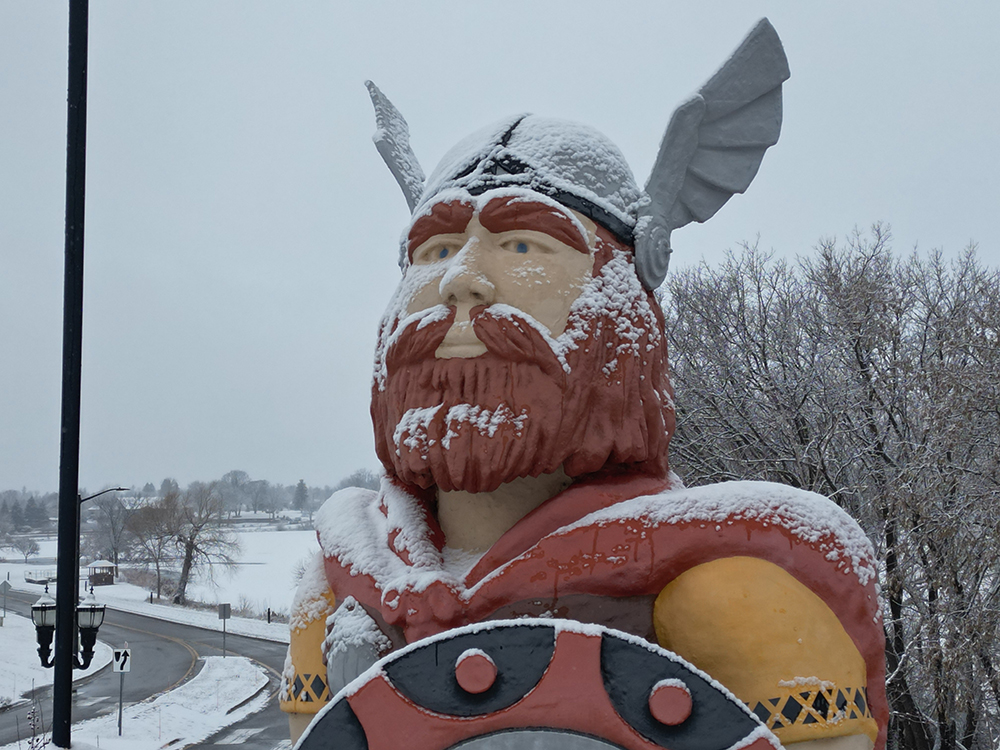 A large Viking statue with a winged helmet and shield is covered in snowfall. The statue stands outdoors near Alexandria, beside a curved road and snowy trees on a cloudy winter day.