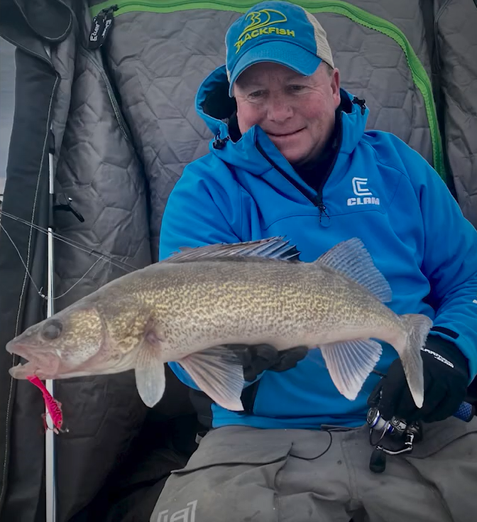A smiling person in a blue jacket and cap holds a large walleye fish inside an ice fishing shelter, likely celebrating success at one of the early ice hot spots, with fishing gear visible in the background.