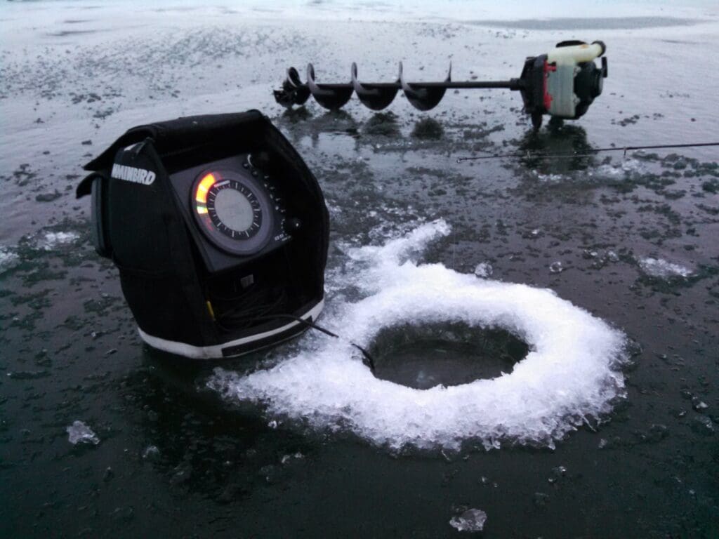 An ice fishing sonar sits next to an icy hole drilled in a frozen lake, helping anglers monitor ice depths in Alexandria area waters, with an ice auger lying nearby on the snowy surface.